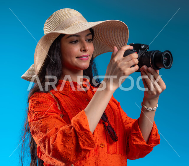 التقاط الصور التذكارية وتوثيق اللحظات السعيدة، الاستمتاع بتصوير الرحلات الصيفية والأماكن السياحية بالمملكة، بورتريه مقرب لشابة عربية خليجية سعودية ترتدي الزي الكاجوال وتمسك كاميرا التصوير الفوتوغرافي، إيماءات تدل على التركيز، خلفية زرقاء.
