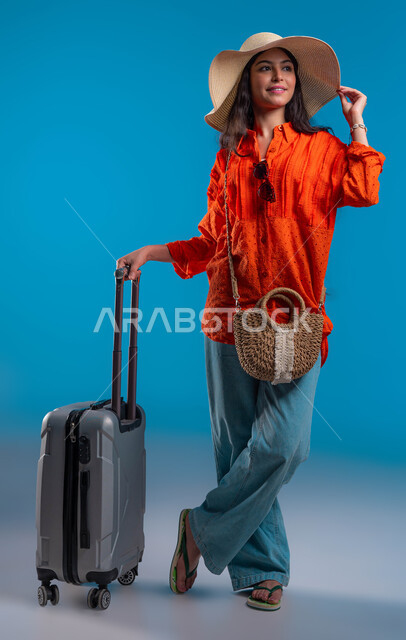 قضاء وقت ممتع مع الأصدقاء والاستمتاع بالعطلة الصيفية, الوقوف بإيماءات الثقة والسعادة والحماس, بورتريه مقرب لامرأة عربية خليجية سعودية ترتدي القبعة والزي الكاجوال وتمسك حقيبة السفر, التجهيز والاستعداد للرحلة السياحية, خلفية زرقاء.