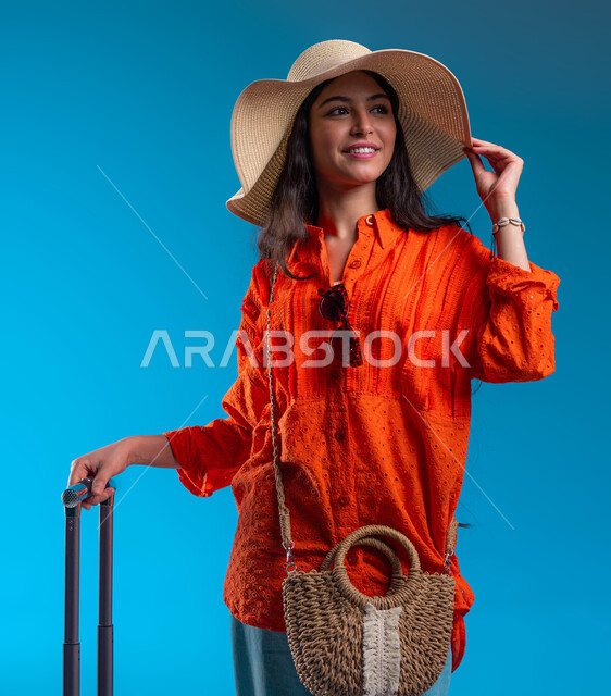 الاستمتاع بالعطلة الصيفية وقضاء وقت ممتع مع الأصدقاء, الوقوف بإيماءات الثقة والسعادة والحماس, بورتريه مقرب لامرأة عربية خليجية سعودية ترتدي القبعة والزي الكاجوال وتمسك حقيبة السفر, التجهيز والاستعداد للرحلة السياحية, خلفية زرقاء.