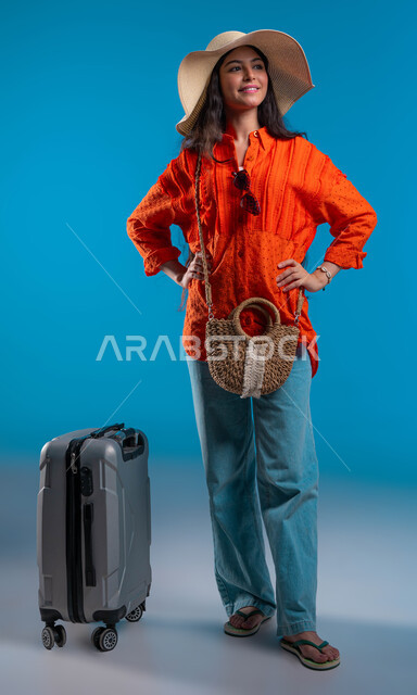 الاستعداد وتجهيز حقيبة السفر لرحلة سياحية سعيدة، الوقوف بإيماءات الثقة والسعادة والسرور, مفهوم الأنوثة والأناقة، بورتريه مقرب لامرأة عربية خليجية سعودية مبتسمة ترتدي القبعة والزي الكاجوال الخاص بالرحلات الصيفية، صورة الجسم بالكامل، خلفية زرقاء.