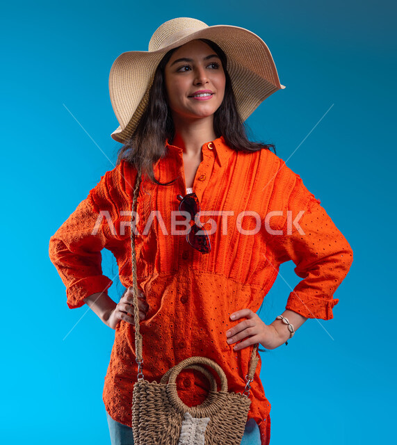 الاهتمام بالمظهر الخارجي, مفهوم الأنوثة والأناقة، بورتريه مقرب لامرأة عربية خليجية سعودية مبتسمة ترتدي القبعة والزي الكاجوال الخاص بالرحلات الصيفية تنظر إلى شيء ما، الوقوف بإيماءات الثقة والسعادة والسرور, خلفية زرقاء.