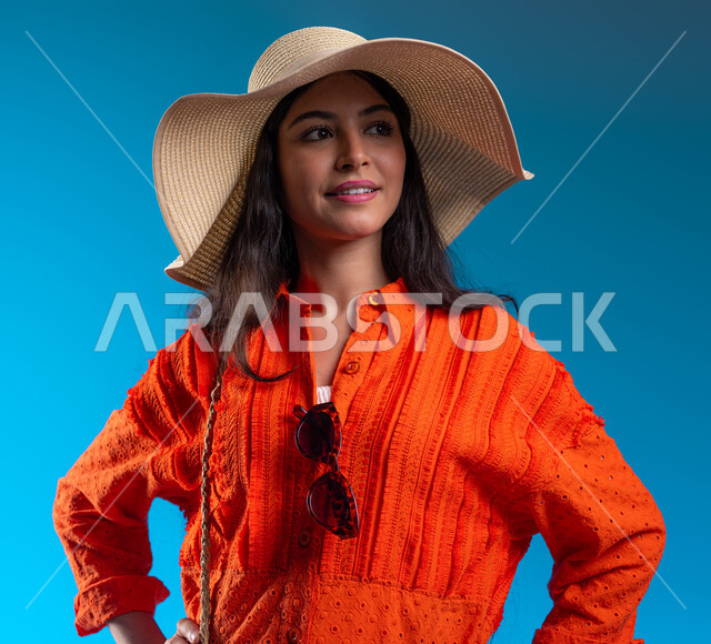 Standing with gestures of confidence, happiness and pleasure, concern for external appearance, concept of femininity and elegance, close-up portrait of a smiling Saudi Arabian Gulf woman wearing a hat and casual clothes for summer trips, looking at something, blue background.