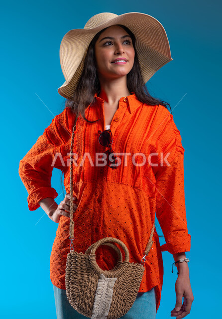 الاهتمام بالمظهر الخارجي, مفهوم الأنوثة والأناقة، بورتريه مقرب لامرأة عربية خليجية سعودية مبتسمة ترتدي القبعة والزي الكاجوال الخاص بالرحلات الصيفية تنظر إلى شيء ما، الوقوف بإيماءات الثقة والسعادة والسرور, خلفية زرقاء.