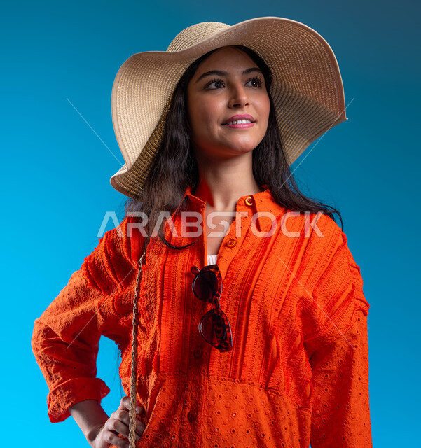 مفهوم الأنوثة والأناقة، الاهتمام بالمظهر الخارجي, بورتريه مقرب لامرأة عربية خليجية سعودية ترتدي القبعة والزي الكاجوال الخاص بالرحلات الصيفية، الوقوف بإيماءات الثقة والسعادة والسرور, خلفية زرقاء.