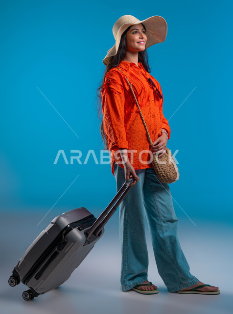الاستمتاع بالعطلة الصيفية وقضاء وقت ممتع مع الأصدقاء, الوقوف بإيماءات الثقة والسعادة والسرور, بورتريه لامرأة عربية خليجية سعودية ترتدي القبعة والزي الكاجوال وتمسك حقيبة السفر, التجهيز والاستعداد للرحلة السياحية, صورة لكامل الجسم، خلفية زرقاء.
