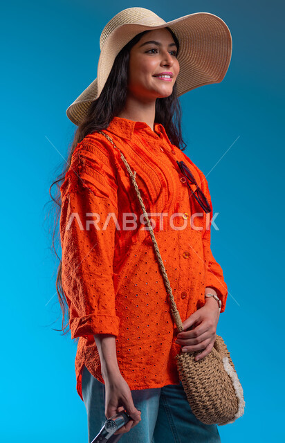 الاستمتاع بالعطلة الصيفية وقضاء وقت ممتع في منتجعات المملكة, بورتريه مقرب لامرأة عربية خليجية سعودية ترتدي القبعة والزي الكاجوال وتمسك حقيبة السفر تقف بإيماءات السعادة والفرح, الاستعداد والتجهيز للرحلة السياحية السعيدة, خلفية زرقاء.