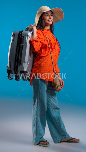 الوقوف بإيماءات الثقة والسعادة والسرور, بورتريه لامرأة عربية خليجية سعودية ترتدي القبعة والزي الكاجوال وتحمل حقيبة السفر, التجهيز والاستعداد للرحلة السياحية, الاستمتاع بالعطلة الصيفية وقضاء وقت ممتع مع الأصدقاء, صورة لكامل الجسم، خلفية زرقاء.