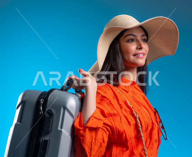 الحماس والاستعداد للرحلة السياحية, الاستمتاع بالعطلة الصيفية وقضاء وقت ممتع مع الأصدقاء, الوقوف بإيماءات الثقة والسعادة والفرح, بورتريه مقرب لامرأة عربية خليجية سعودية ترتدي القبعة والزي الكاجوال وتحمل حقيبة السفر, خلفية زرقاء.