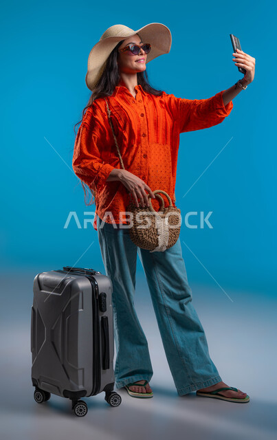 توثيق اللحظات السياحية الممتعه, بورتريه لشابة عربية خليجية سعودية مبتسمة ترتدي الزي الكاجوال والنظارة الشمسية مع قبعة القش تقوم بالتقاط صور سيلفي تذكارية عبر الهاتف المحمول, السفر والاستمتاع بالأجواء الصيفيه, صورة الجسم بالكامل، خلفية زرقاء