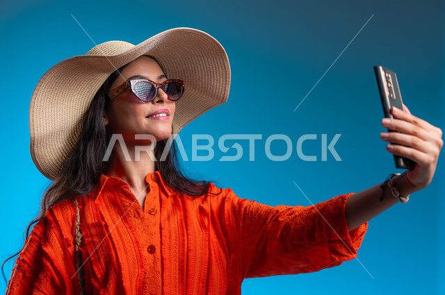 توثيق اللحظات السياحية الممتعه باستخدام الجوال, الاهتمام بالأزياء والموضة العصرية، بورتريه مقرب لشابة عربية خليجية سعودية مبتسمة ترتدي الزي الكاجوال والنظارة الشمسية مع قبعة القش الصيفية تقوم بالتقاط صور سيلفي تذكارية عبر الهاتف المحمول، خلفية زرقاء.