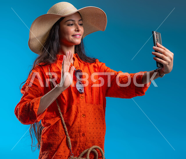 الاستمتاع والتسلية والترفيه بالعطلة الصيفية، بورتريه مقرب رلامرأة عربية خليجية سعودية ترتدي الزي الكاجوال والقبعة تجري مكالمة فيديو عبر الهاتف المحمول ترفع يدها بإيماءات تدل على إلقاء التحية، مفهزوم الأنوثة والأناقة، خلفية زرقاء.
