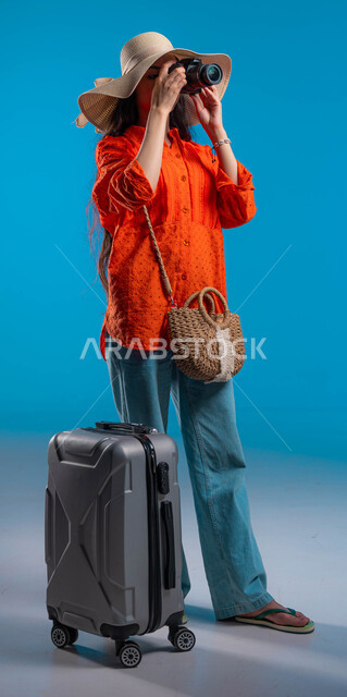 التقاط الصور التذكارية وتوثيق اللحظات السعيدة، الاستمتاع بتصوير الأماكن السياحية بالمملكة، بورتريه لشابة عربية خليجية سعودية ترتدي الزي الكاجوال وتستخدم كاميرا التصوير الفوتوغرافي، تجهيز حقيبة السفر لقضاء إجازة صيفية ممتعة، صورة الجسم بالكامل، خلفية زرقاء.
