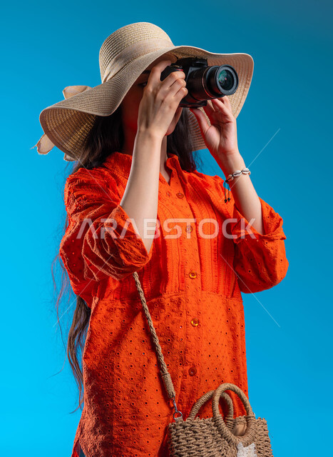 الاستمتاع بتصوير الرحلة والأماكن السياحية بالمملكة، استخدام كاميرا التصوير الفوتوغرافي لالتقاط الصور التذكارية وتوثيق اللحظات السعيدة، بورتريه مقرب لشابة عربية خليجية سعودية ترتدي لباس كاجوال خاص بالرحلات الصيفية، إيماءات تدل على التركيز، خلفية زرقاء.