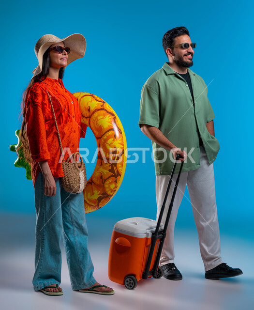 أجواء عائلية وصيفية ممتعة، استخدام حافظة التبريد للاستمتاع بالمشروبات الباردة، بورتريه لزوجين عربيين خليجيين سعوديين مبتسمين يرتديان الزي الكاجوال والنظارة الشمسية، السباحة في العوامة في شواطئ السعودية بإيماءات السرور, صورة الجسم بالكامل، خلفية زرقاء