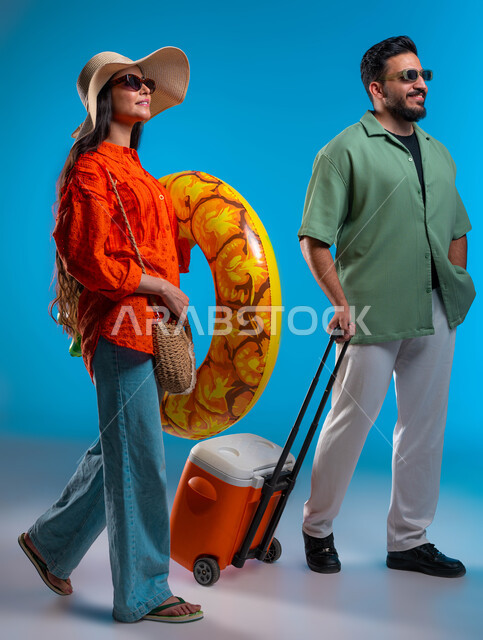 استخدام حافظة التبريد للاستمتاع بالمشروبات الباردة، قضاء العطلة الصيفية مع العائلة, بورتريه لزوجين عربيين خليجيين سعوديين مبتسمين يرتديان الزي الكاجوال والنظارة الشمسية، السباحة في العوامة في شواطئ السعودية بإيماءات السرور, صورة الجسم بالكامل، خلفية زرقاء