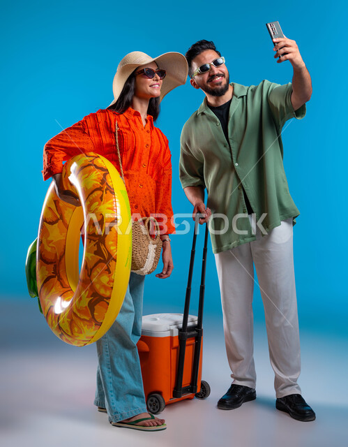 التقاط صور سيلفي تذكارية عبر الهاتف المحمول, الاستمتاع بالمشروبات الباردة وقضاء وقت ممتع في شواطئ السعودية,بورتريه لشاب وشابة عربين خليجيين سعوديين مبتسمين يرتديان الزي الكاجوال يقومان بتوثيق اللحظات السعيدة بإيماءات السرور,  صورة الجسم بالكامل,  خلفية زرقاء