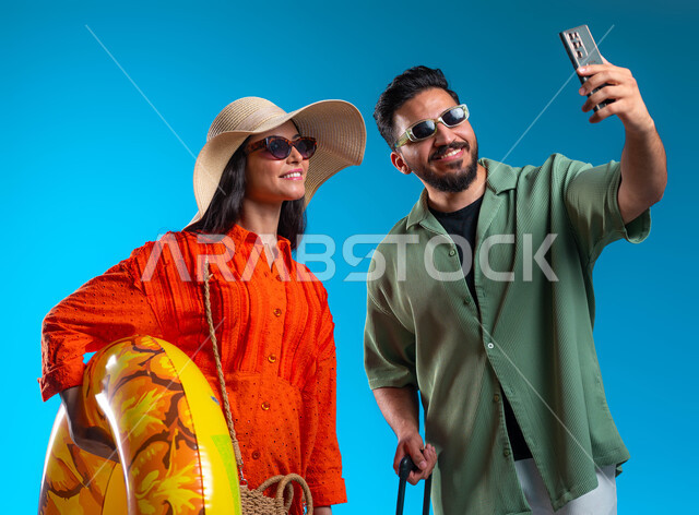 التقاط صور سيلفي تذكارية عبر الهاتف المحمول, قضاء وقت ممتع في شواطئ السعودية,بورتريه مقرب لشاب وشابة عربين خليجيين سعوديين مبتسمين يرتديان الزي الكاجوال يقومان بتوثيق اللحظات السعيدة بإيماءات السرور والسعادة,  خلفية زرقاء