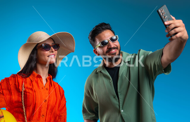 استخدام الهاتف المحمول لالتقاط صور السيلفي التذكارية, قضاء وقت ممتع في شواطئ السعودية,بورتريه مقرب لشاب وشابة عربين خليجيين سعوديين مبتسمين يرتديان الزي الكاجوال يقومان بتوثيق اللحظات السعيدة بإيماءات السرور والسعادة,  خلفية زرقاء