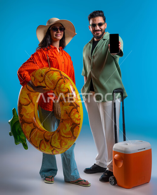 عرض تطبيقات وخدمات السياحة والسفر, الاستمتاع بالمشروبات الباردة وقضاء وقت ممتع في شواطئ السعودية,بورتريه مقرب لشاب وشابة عربين خليجيين سعوديين مبتسمين يرتديان الزي الكاجوال والنظارة الشمسية, شابة سعودية تحمل بيدها عوامة السباحة، صورة الجسم بالكامل، خلفية زرقاء
