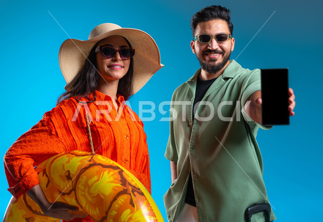 قضاء وقت ممتع في شواطئ السعودية, عرض تطبيقات وخدمات السياحة والسفر, بورتريه مقرب لشاب وشابة عربين خليجيين سعوديين مبتسمين يرتديان الزي الكاجوال والنظارة الشمسية, شابة سعودية تحمل بيدها عوامة السباحة، ​خلفية زرقاء