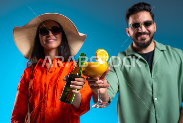 النظر إلى الكاميرا بإيماءات السرور والسعادة، أجواء عائلية سعيدة, بورتريه مقرب لرجل وامرأة عربيين خليجين سعوديين مبتسمين يرتديان الزي الكاجوال والنظارات الشمسية يقومان بشرب العصائر المنعشة, الاستمتاع بالمشروبات الباردة في الأجواء الحارة، خلفية زرقاء