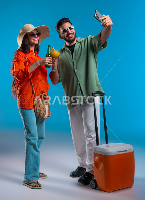 الاستمتاع بالمشروبات الباردة الصيفيه,  ارتداء الزي الكاجوال والنظارات الشمسية, إيماءات السرور والتقاط صور سليفي تذكارية عبر الجوال, بورتريه مقرب لشاب وشابة عربين خليجيين سعوديين مبتسمان يقومان بتوثيق اللحظات السعيدة, صورة الجسم بالكامل, خلفية زرقاء