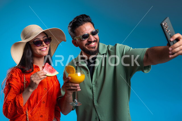 Taking souvenir selfies via mobile phone, a close-up portrait of a smiling young Saudi Gulf Arab man and woman, wearing casual clothes, documenting happy moments with gestures of joy and happiness, enjoying drinks and summer fruits, entertainment, amusement, and spending a pleasant vacation, blue background.
