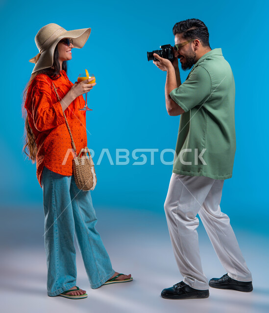 توثيق اللحظات السعيدة, مصور سعودي يقوم بالتقاط صور فوتوغرافية باستخدام كاميرا عالية الدقة, بورتريه مقرب من الجانب لشابة عربية خليجية سعودية ترتدي الزي الكاجوال وقبعة من القش تمسك كوب العصير, الاستمتاع بالإجازة الصيفية, صورة الجسم بالطول الكامل,  خلفية زرقاء