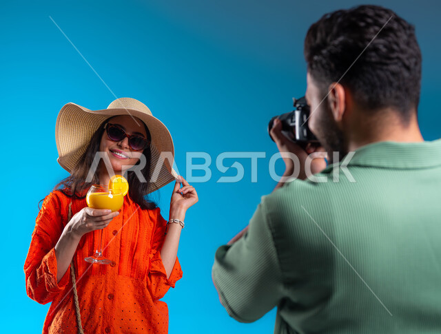 الأجواء الصيفية والاستمتاع بالرحلة السياحية, قضاء وقت ممتع مع العائلة, بورتريه مقرب من الخلف لشاب عربي خليجي سعودي يرتدي الزي الكاجوال يقوم بالتقاط صور فوتوغرافية باستخدام كاميرا عالية الدقة, شابة سعودية تقوم بشرب العصير, خلفية زرقاء