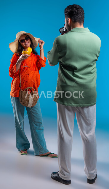 قضاء وقت ممتع مع العائلة, بورتريه من الخلف لشاب عربي خليجي سعودي يرتدي الزي الكاجوال يقوم بالتقاط صور فوتوغرافية باستخدام  كاميرا عالية الدقة, شابة سعودية تقوم بشرب العصير, الاستمتاع بالرحلة السياحية والأجواء الصيفية, صورة الجسم بالطول الكامل, خلفية زرقاء