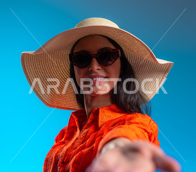 Entertainment, entertainment and enjoying the summer vacation, a close-up portrait of a smiling Saudi Arabian Gulf woman wearing a casual outfit and a straw hat looking at the camera, cheerfulness and pleasure with marriage proposal gestures, a pleasant tourist trip, blue background