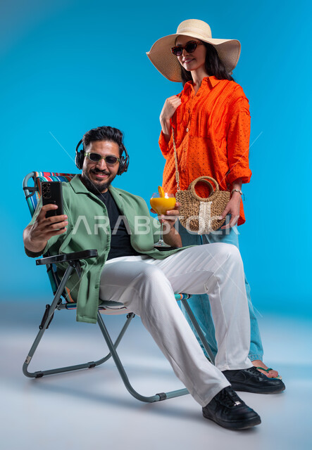 الاستمتاع وقضاء وقت ممتع في إحدى الرحلات السياحية, ارتداء النظارات الشمسية والأزياء الصيفية المريحة, التقاط صور سيلفي تذكارية, بورتريه لشاب وشابة عربيين خليجيين سعوديين يقومان بتوثيق اللحظات السعيدة، صورة الجسم بالطول الكامل, خلفية زرقاء