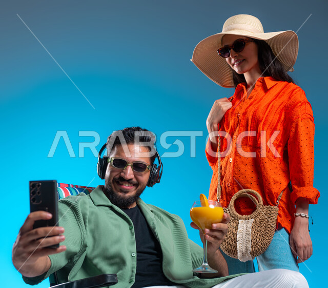 ارتداء النظارات الشمسية والأزياء الصيفية المريحة, التقاط صور سيلفي تذكارية, بورتريه لشاب وشابة عربيين خليجيين سعوديين يقومان بتوثيق اللحظات السعيدة، الاستمتاع وقضاء وقت ممتع في إحدى الرحلات السياحية، خلفية زرقاء