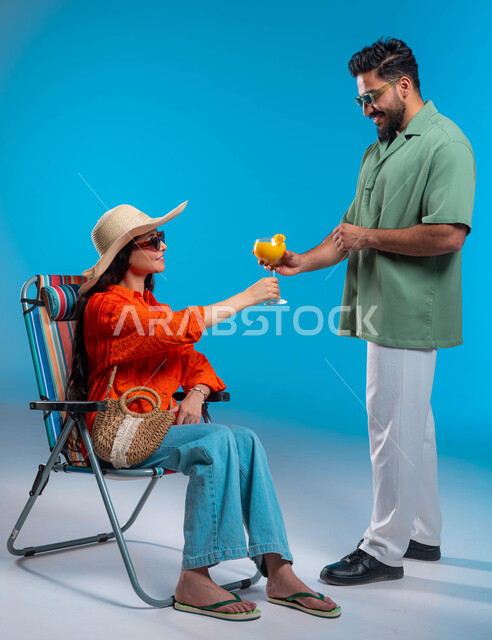 Recreation, tourism and spending enjoyable summer times, a portrait of a smiling Saudi Gulf Arab woman wearing a casual outfit and a straw hat enjoying a tourist trip, a Saudi man wearing sunglasses serving refreshing natural juice, full-length body portrait, blue background