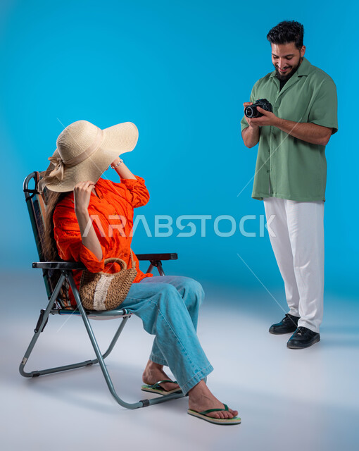 توثيق اللحظات السعيدة والتقاط الصور التذكارية, بورتريه لشاب عربي خليجي سعودي يرتدي الزي الكاجوال يقوم باستخدام كاميرا فوتوغرافية عالية الدقة, شابة سعودية تستمتع بجلسة تصوير في الرحلة السياحية, قضاء أوقات ممتعه بالعطلة الصيفية, صورة الجسم بالكامل, خلفية زرقاء