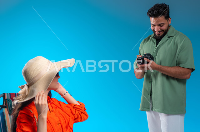 Taking souvenir photos, a portrait of a young Saudi Gulf Arab man wearing a casual outfit using a high-resolution photographic camera, a young Saudi woman wearing a straw hat enjoying a photo session in one of the tourist areas, spending fun times during the summer vacation, blue background