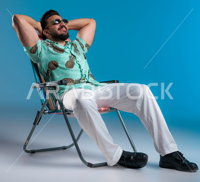 الجلوس والاستمتاع بالإجازة والمناظر الطبيعيه, الرحلات السياحية في إحدى المنتجعات والشواطئ السعودية, بورتريه لشاب عربي خليجي سعودي مبتسم يرتدي الزي الكاجوال والنظارة يرفع يده للأعلى, النظر إلى شيء ما بإيماءات الاسترخاء والراحة, خلفية زرقاء