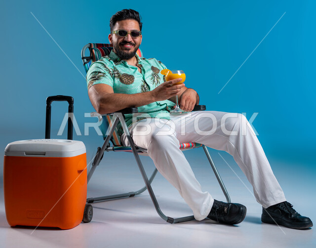 قضاء إجازة صيفية ممتعه, النظر إلى الكاميرا بإيماءات السرور, شرب العصائر الطبيعيه المنعشة, بورتريه لشاب عربي خليجي سعودي مبتسم يرتدي الزي الكاجوال والنظارة الشمسية يستمتع في إحدى الشواطئ والمنتجعات الترفيهيه, السياحة والاصطياف في السعودية, خلفية زرقاء