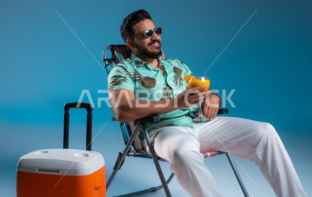 النظر إلى شيء ما بإيماءات السرور, شرب العصائر الطبيعيه المنعشة, بورتريه لشاب عربي خليجي سعودي مبتسم يرتدي الزي الكاجوال والنظارة الشمسية يستمتع في إحدى الشواطئ والمنتجعات الترفيهيه, السياحة والاصطياف في السعودية, قضاء إجازة صيفية ممتعه, خلفية زرقاء