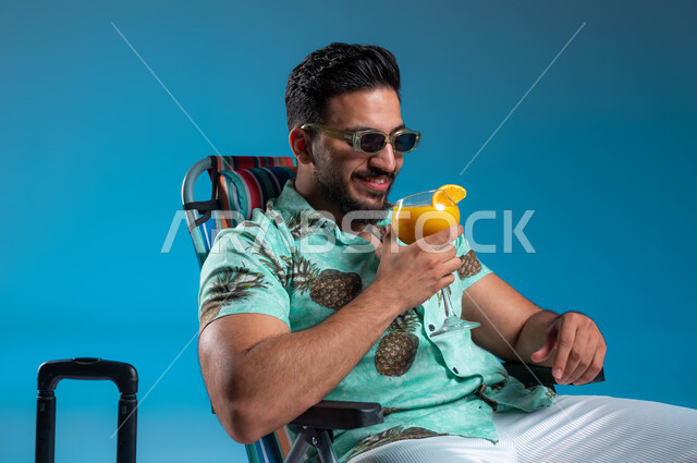 شرب العصائر الطبيعيه المنعشة, بورتريه مقرب لشاب عربي خليجي سعودي مبتسم يرتدي الزي الكاجوال والنظارة الشمسية يجلس على الكرسي في إحدى الشواطئ والمنتجعات الترفيهيه, السياحة والاصطياف في السعودية, الاستمتاع بالاجازة الصيفية, خلفية زرقاء
