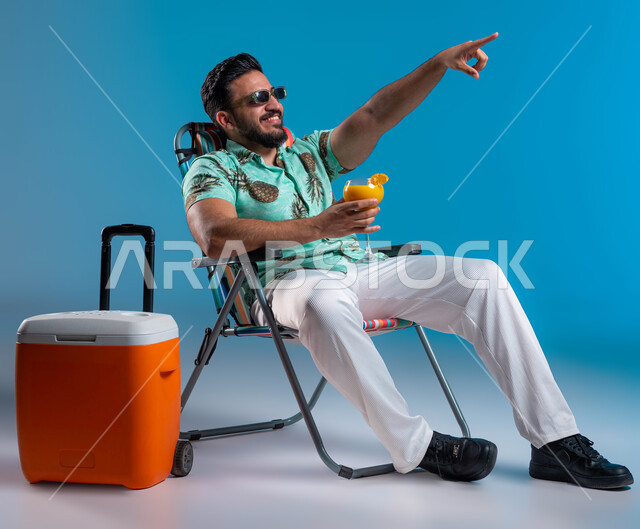 الاشارة إلى شيء ما والاستمتاع بالجلسات االهادئة, بورتريه لشاب عربي خليجي سعودي مبتسم يرتدي الزي الكاجوال والنظارة الشمسية يجلس على الكرسي في إحدى الشواطئ والمنتجعات الترفيهيه, السياحة والاصطياف في دول الخليج, شرب العصائر الطبيعيه المنعشة, خلفية زرقاء
