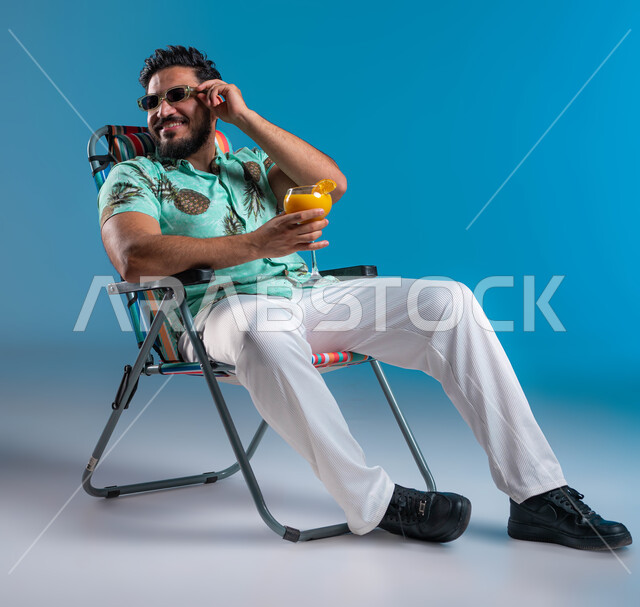 شرب العصائر الطبيعيه المنعشة, قضاء أوقات سياحية ممتعه, الاستمتاع بالجلسات االهادئة والنظر إلى شيء ما, بورتريه لشاب عربي خليجي سعودي مبتسم يرتدي الزي الكاجوال والنظارة الشمسية يجلس على الكرسي في إحدى الشواطئ والمنتجعات الترفيهيه, خلفية زرقاء