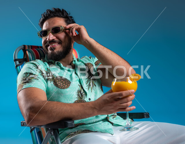 الاستمتاع بالجلسات االهادئة والنظر إلى شيء ما, بورتريه مقرب لشاب عربي خليجي سعودي مبتسم يرتدي الزي الكاجوال والنظارة الشمسية يجلس على الكرسي في إحدى الشواطئ والمتتجعات الترفيهيه, قضاء أوقات سياحية ممتعه, شرب العصائر الطبيعيه المنعشة, خلفية زرقاء