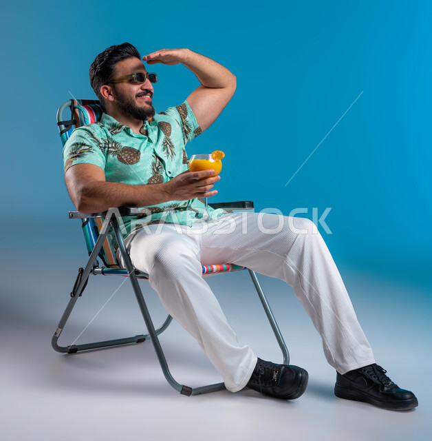 النظر إلى شيء ما والاستمتاع بالجلسات االهادئة, بورتريه لشاب عربي خليجي سعودي مبتسم يرتدي الزي الكاجوال والنظارة الشمسية يجلس على الكرسي في إحدى الشواطئ والمتتجعات الترفيهيه, رفع اليد للأعلى والحماية من أشعة الشمس, شرب العصائر الطبيعيه المنعشة, خلفية زرقاء