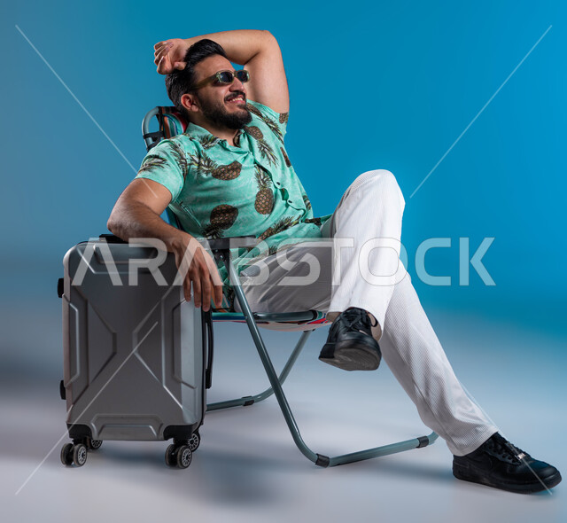 الاستمتاع بالإجازة والمناظر الطبيعيه, الارتكاز على حقيبة السفر والنظر إلى شيء ما, الرحلات السياحية في إحدى المنتجعات والشواطئ السعودية, بورتريه لشاب عربي خليجي سعودي مبتسم يرتدي الزي الكاجوال والنظارة يرفع يده للأعلى بإيماءات الاسترخاء والراحة, خلفية زرقاء