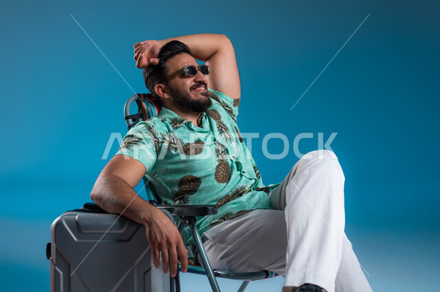 الارتكاز على حقيبة السفر والنظر إلى شيء ما, الرحلات السياحية في إحدى المنتجعات والشواطئ السعودية, بورتريه لشاب عربي خليجي سعودي مبتسم يرتدي الزي الكاجوال والنظارة يرفع يده للأعلى بإيماءات الاسترخاء والراحة, الاستمتاع بالإجازة والمناظر الطبيعيه, خلفية زرقاء