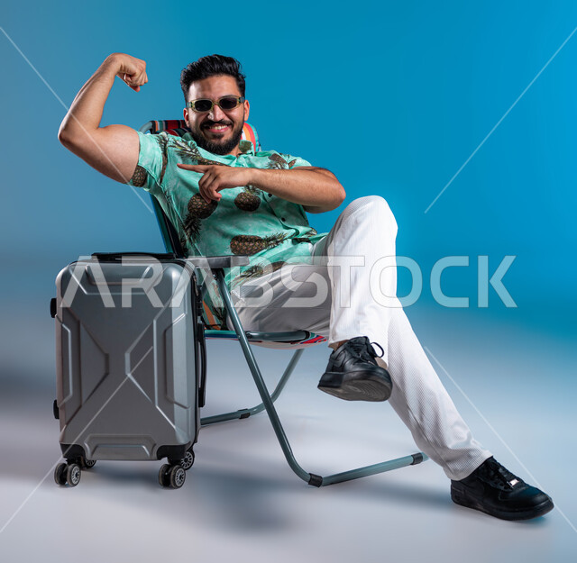 الجسم الصحي وعرض العضلات بإيماءات الثقة والسرور, استعراض القوة البدنية, بورتريه لشاب عربي خليجي سعودي مبتسم يرتدي الزي الكاجوال والنظارة الشمسية يرفع يده بكفة مقبوضة, أهمية السفر وقضاء أوقات ممتعة في الأماكن السياحية, خلفية زرقاء