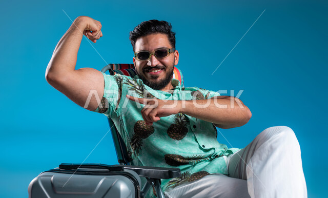 Show of physical strength, portrait of a smiling Saudi Gulf Arab young man wearing casual clothes and sunglasses raising his hand with a clenched palm, healthy body and display of muscles with gestures of confidence and pleasure, the importance of traveling and spending enjoyable times in tourist places, blue background