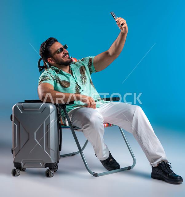 توثيق اللحظات السياحية الممتعه, التواصل مع الأصدقاء والقيام بالبثوب المباشرة عبر الجوال, بورتريه لشاب عربي خليجي سعودي مبتسم يرتدي الزي الكاجوال والنظارة يقوم بالتقام صور سيلفي تذكارية عبر الهاتف المحمول, السفر والاستمتاع بالأجواء الصيفيه, خلفية زرقاء