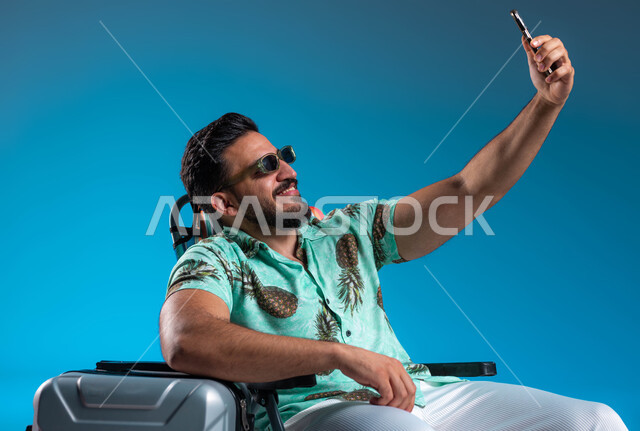 التواصل مع الأصدقاء والقيام بالبثوب المباشرة عبر الجوال, بورتريه لشاب عربي خليجي سعودي مبتسم يرتدي الزي الكاجوال والنظارة يقوم بالتقام صور سيلفي تذكارية عبر الهاتف المحمول, توثيق اللحظات السياحية الممتعه, السفر والاستمتاع بالأجواء الصيفيه, خلفية زرقاء